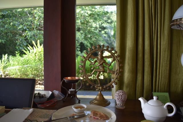 various iteams including a tea pot plate, Shiva Nataraja statue, ipad on a desk, overlooking a window to a green garden