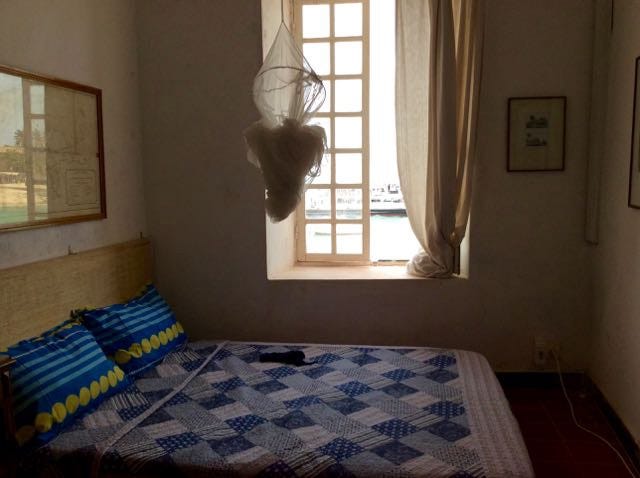 picture of a bed with a blue and white quilty. bundled mosquito net is hanging above, there is an open window overlooking the sea
